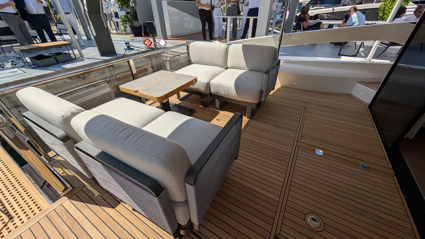 Lounge area on a yacht featuring grey sofas and a wooden coffee table on a teak deck with people visible in the background.